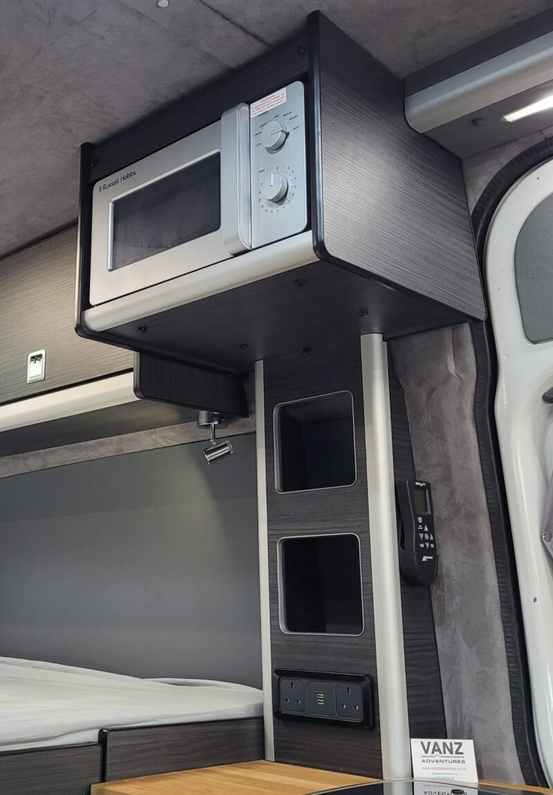 A microwave in its overhead shelf, high on the wall beside a bed